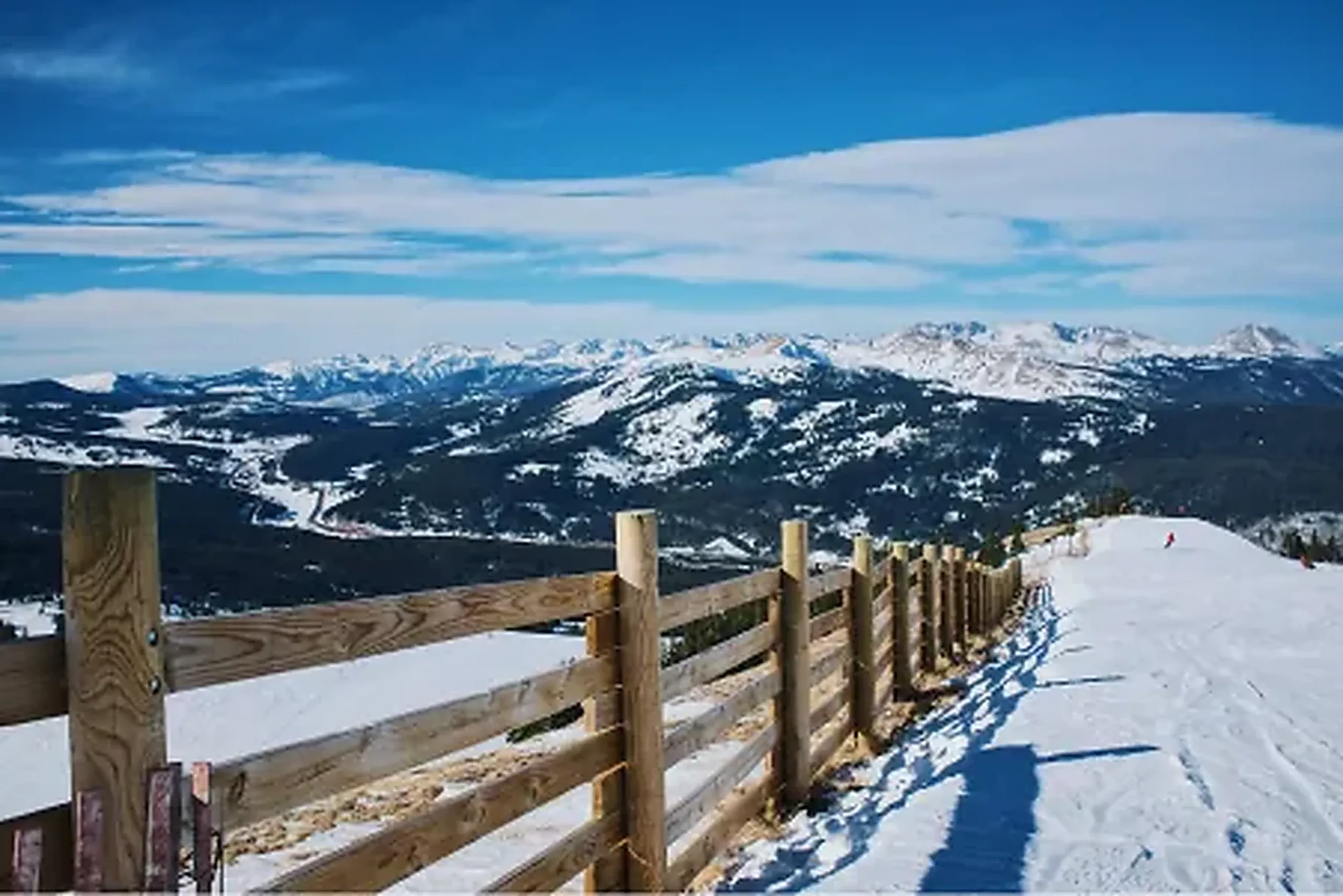 Groomed Colorado ski run beside a snow fence with high peaks in the distance